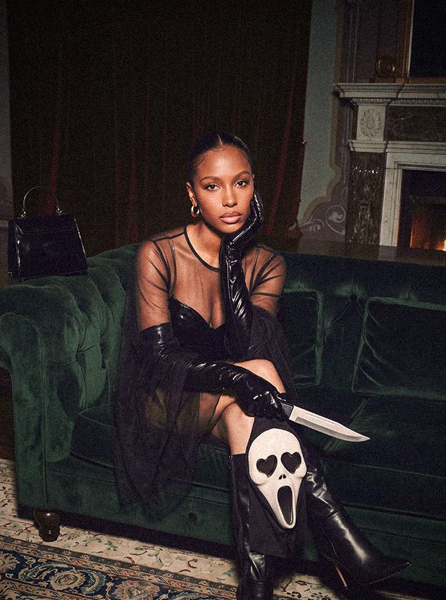 Woman in a chic black mesh Halloween outfit sits on a green velvet sofa holding a knife and Scream mask, showcasing spooky party style.