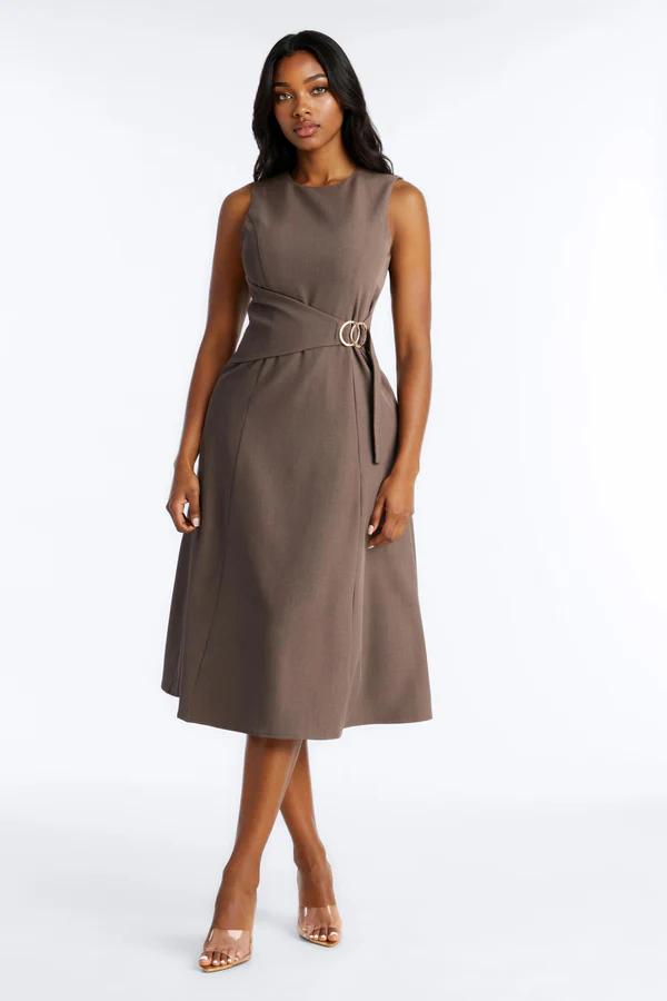 Woman in a sleeveless taupe midi dress featuring a distinctive waist design with a gold buckle, standing on a white background with clear heels.