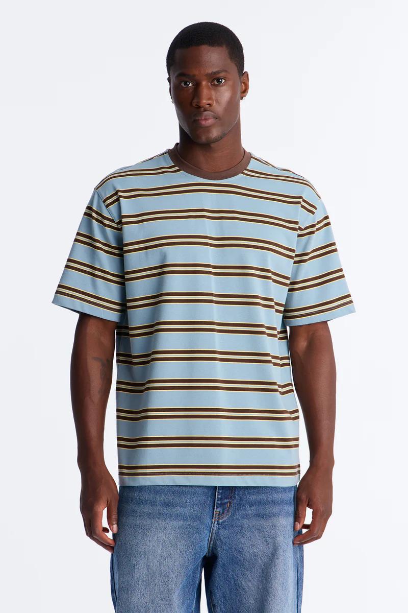 Man models a relaxed-fit light blue, brown, and beige striped men's t-shirt with light wash jeans. Perfect casual wear for the new collection.