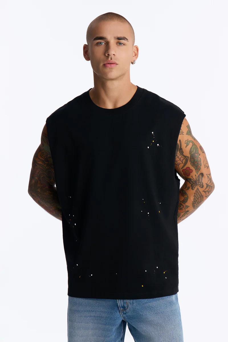 Man models stylish black sleeveless top with white/yellow paint splatter and blue jeans. Discover men's casual wear.