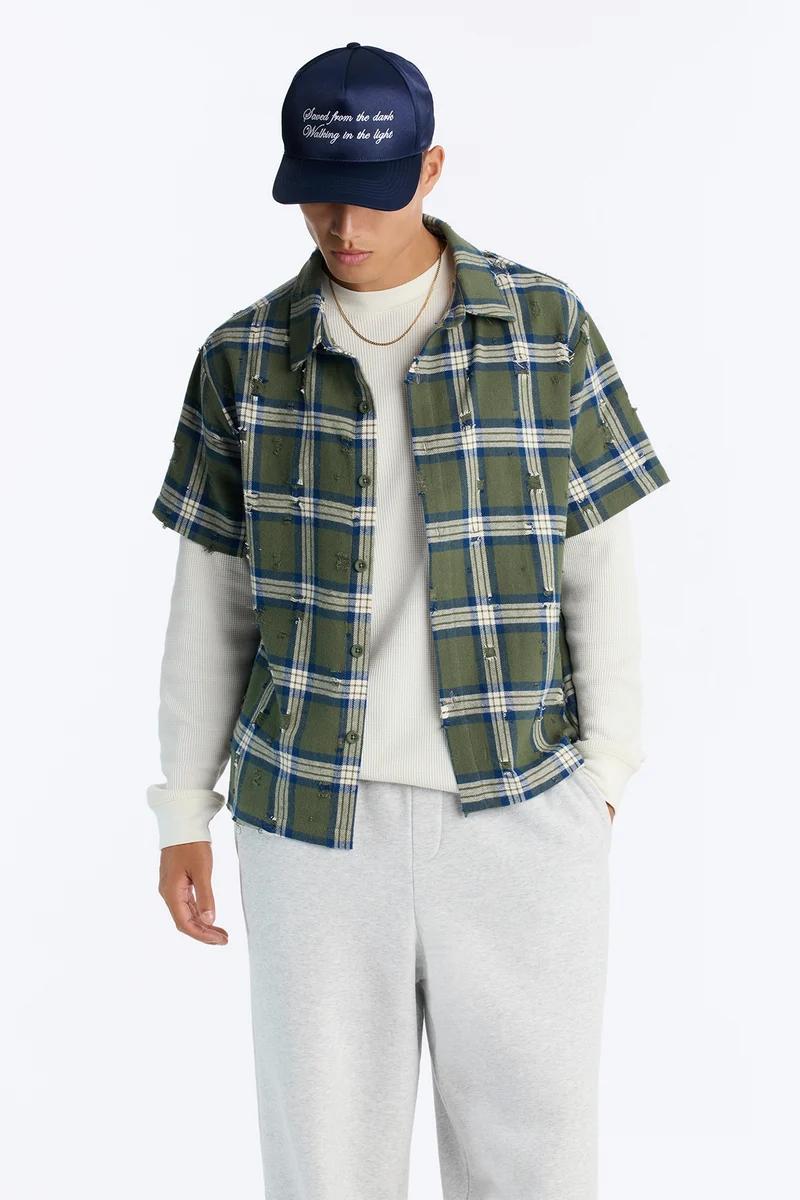 Man wears navy baseball cap, olive distressed plaid short-sleeve shirt, cream long-sleeve tee, grey sweatpants, gold chain. Shop men's casual streetwear.