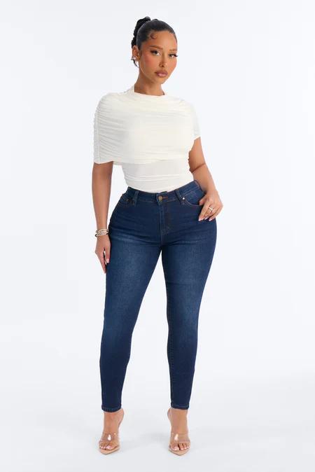 Woman standing, wearing a cream ruched short-sleeve top, dark blue skinny jeans, and clear high-heeled mules on a white background.