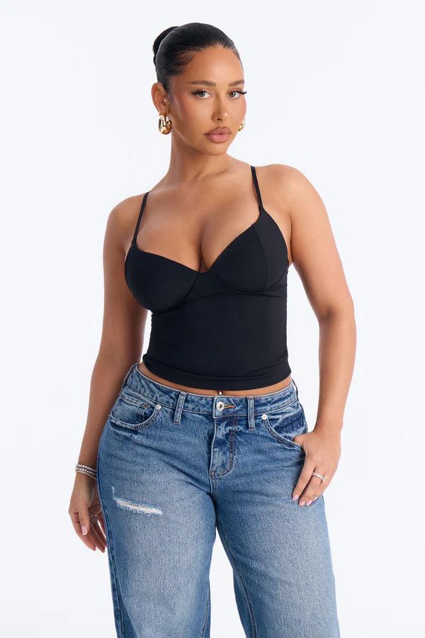 Woman with hair pulled back in a bun, wearing a black strappy bustier top, distressed blue jeans, and gold hoop earrings on a white background.