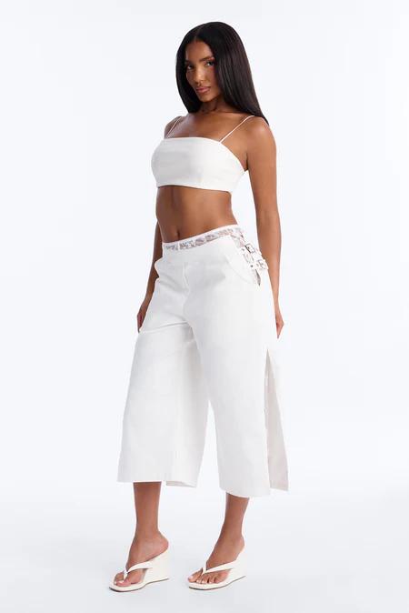 Mujer con top blanco de tirantes y pantalones anchos blancos con encaje lateral y aberturas, llevando chanclas de cuña.
