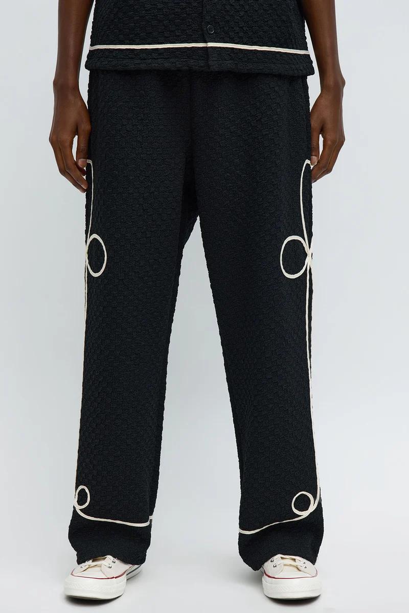 Pantalones negros arrugados con lazos de ribete blanco en laterales y bajo, con top a juego, mostrando la nueva moda masculina.
