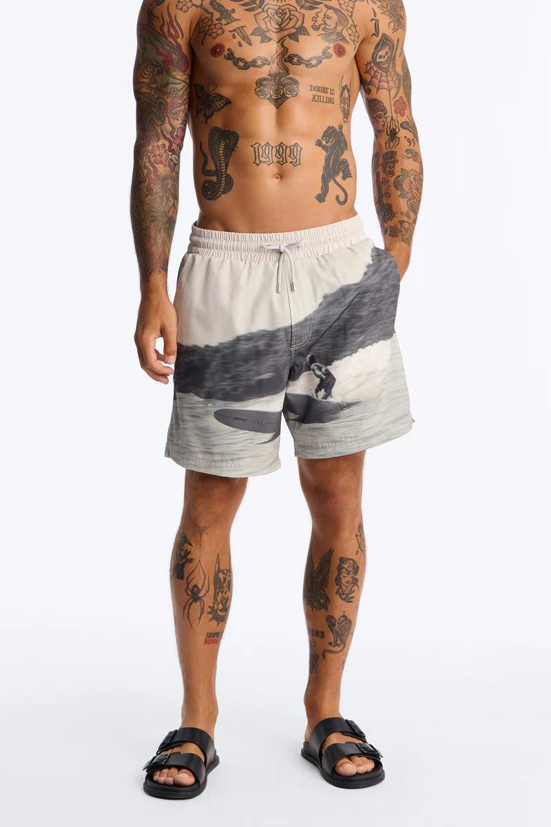 Man wears light grey and black surfing print board shorts with black sandals, promoting versatile men's swimwear.