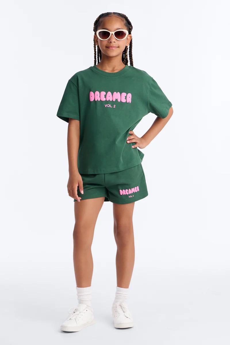 Niña en conjunto verde "DREAMER VOL. 2" de camiseta y shorts, gafas de sol blancas, pelo trenzado. Moda casual infantil.