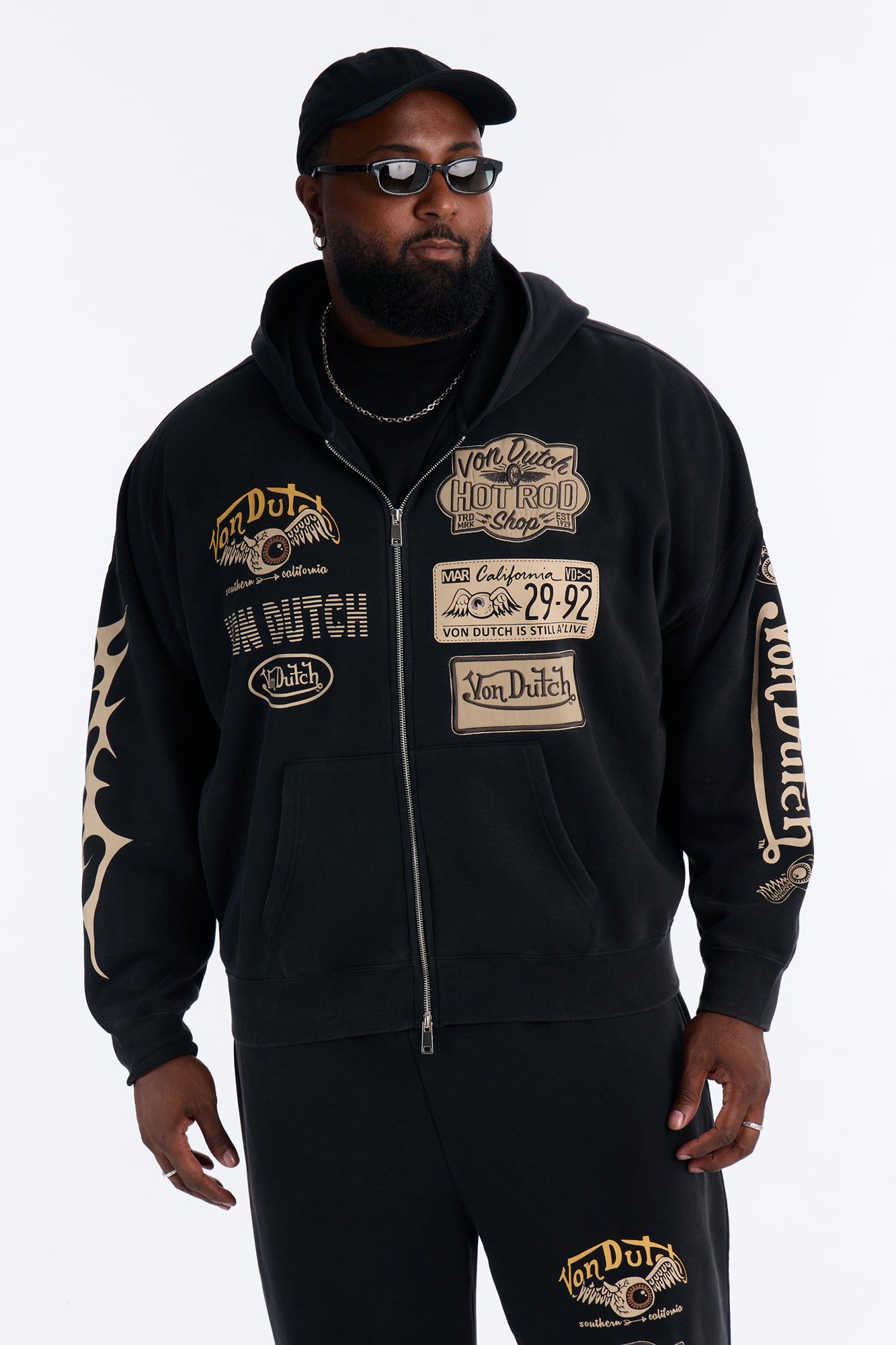 Man wearing black men's Von Dutch patchwork zip-up hoodie with tan graphic logos, sleeve flames. Highlighting new streetwear.