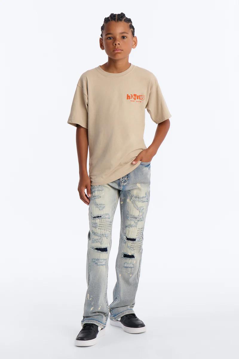 Boy in beige "HATER'S" graphic tee, distressed light wash jeans with patches, and black sneakers. Youth casual fashion.
