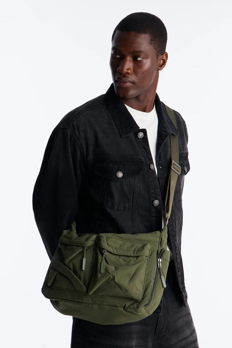 Man wears black denim jacket, white tee, and stylish olive green utility messenger bag with multiple pockets, showcasing versatile men's fashion.