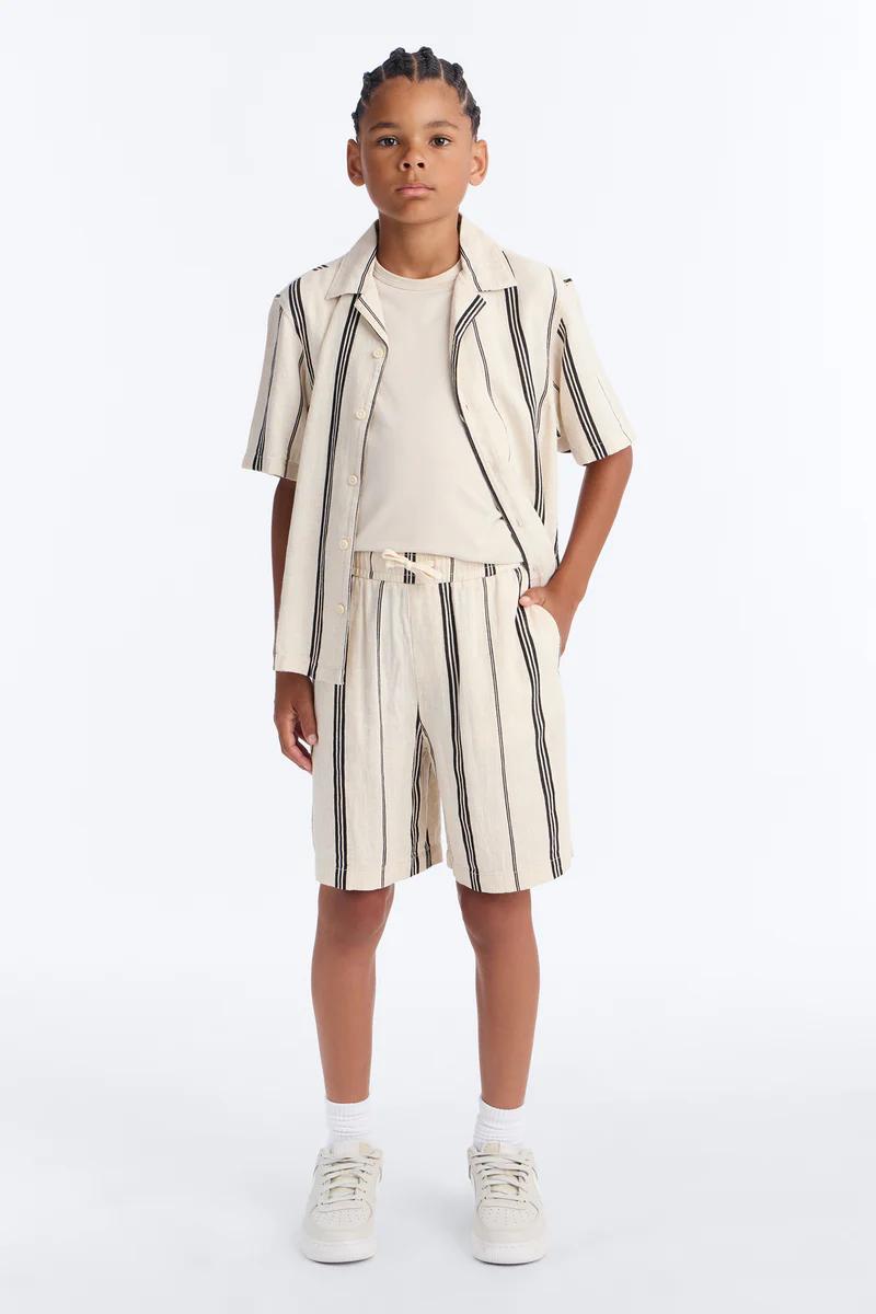 Niño con camisa resort de rayas, shorts a juego y zapatillas. Moda casual de verano para niños, beige con rayas negras.