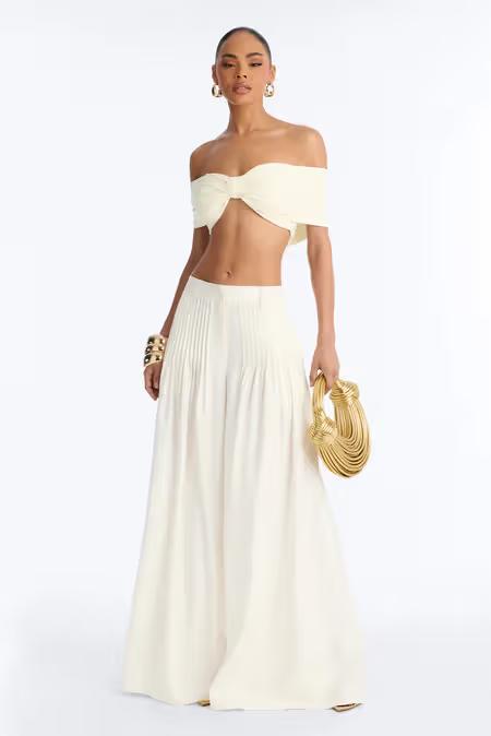 Mujer elegante con top crema escote Bardot, pantalones plisados anchos, accesorios dorados y bolso único.