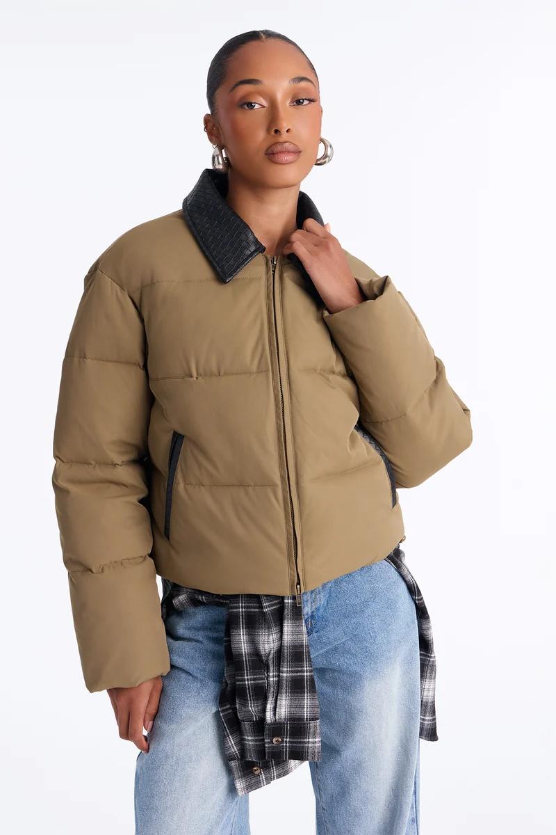Woman wearing a tan cropped puffer jacket with a black leather collar, blue jeans, and a flannel shirt.