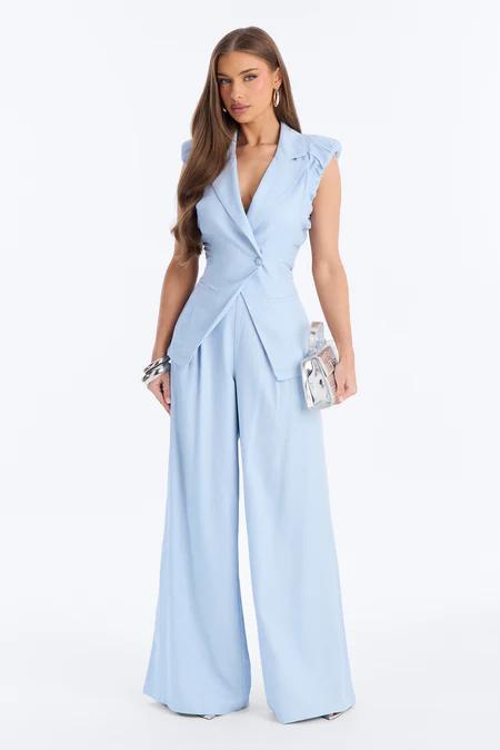 Woman in a light blue sleeveless blazer jumpsuit with wide-leg pants, holding a silver clutch, standing against a white background.