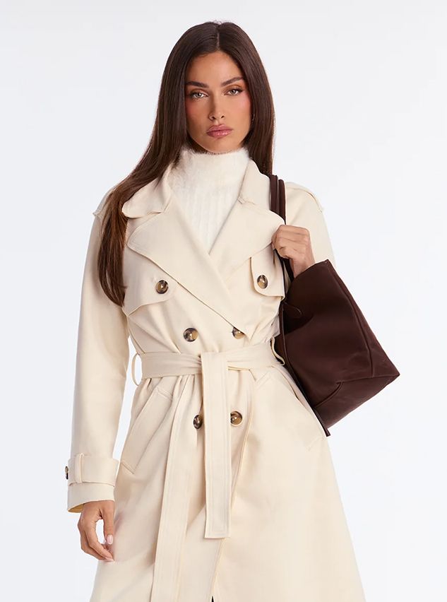 Woman wearing a chic cream double-breasted trench coat and white sweater, carrying a brown tote bag. Fall women's outerwear fashion collection.