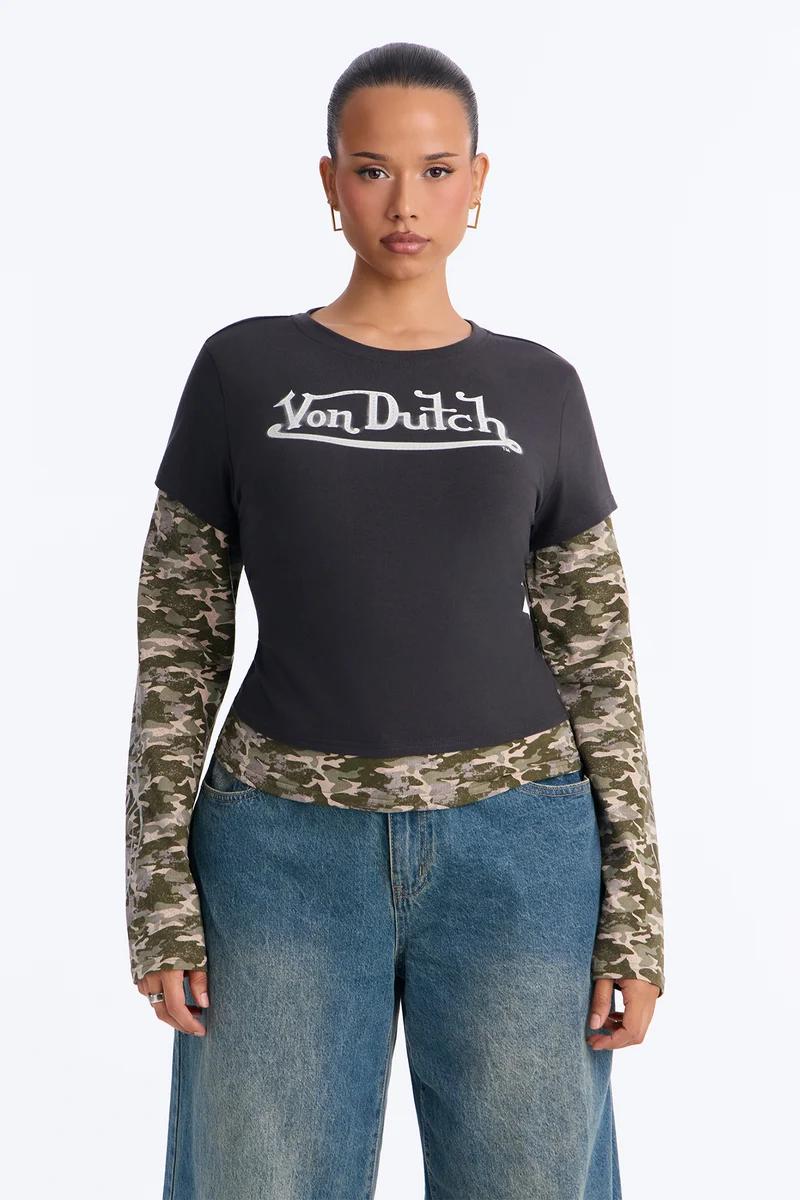Model wearing a black Von Dutch logo graphic top with layered camo long sleeves and blue jeans.