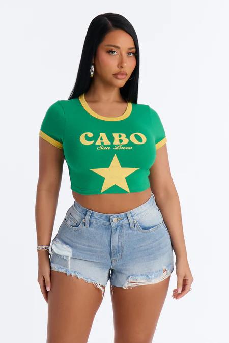 Mujer de pie con top corto verde y amarillo "CABO San Lucas", shorts de mezclilla azules rasgados y pendientes de aro plateados.
