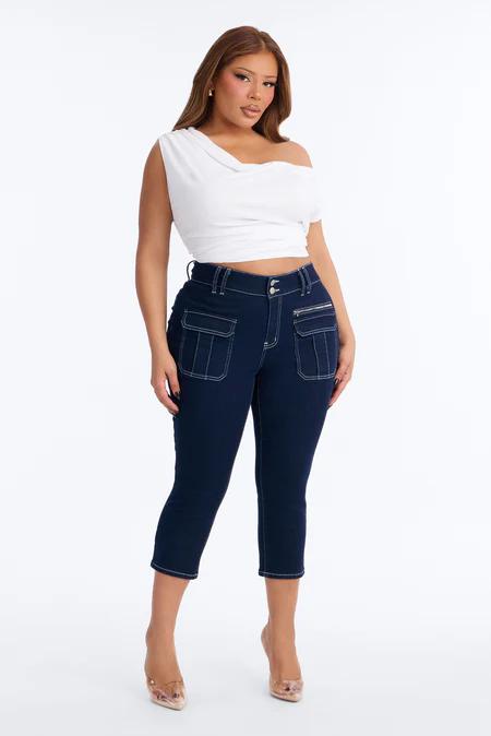 Mujer con top blanco de un hombro y vaqueros capri cargo azul marino oscuro con pespuntes blancos, de pie sobre fondo blanco.