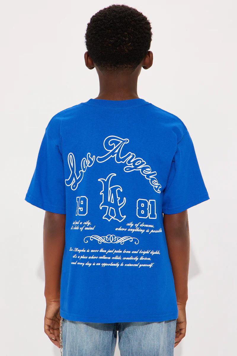 Smiling boy in blue Los Angeles 1981 graphic tee, light wash jeans with chain, & white sneakers. Kids fashion.