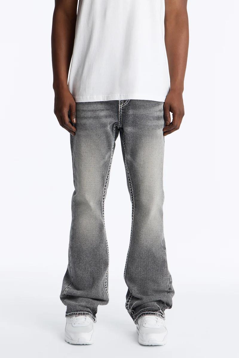 Worn-in grey bootcut jeans with white contrast stitching worn by a person in a white tee and sneakers. Shop men's denim.