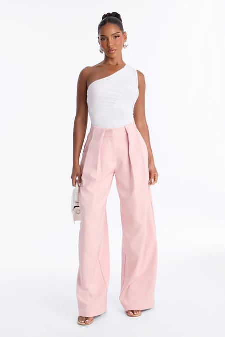Vista de cuerpo completo de una mujer con top blanco de un hombro, pantalones anchos plisados rosa claro, sandalias nude y bolso blanco.