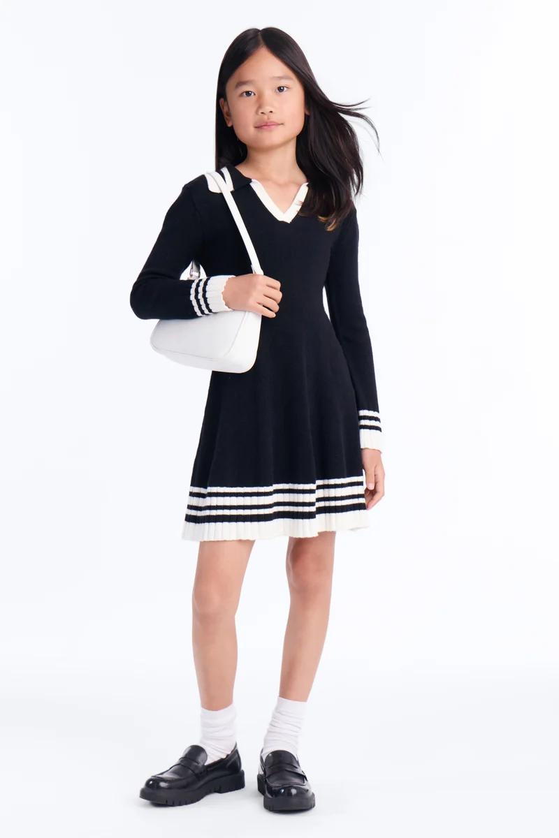 Niña con vestido de punto negro de manga larga con rayas blancas, bolso blanco y mocasines negros.