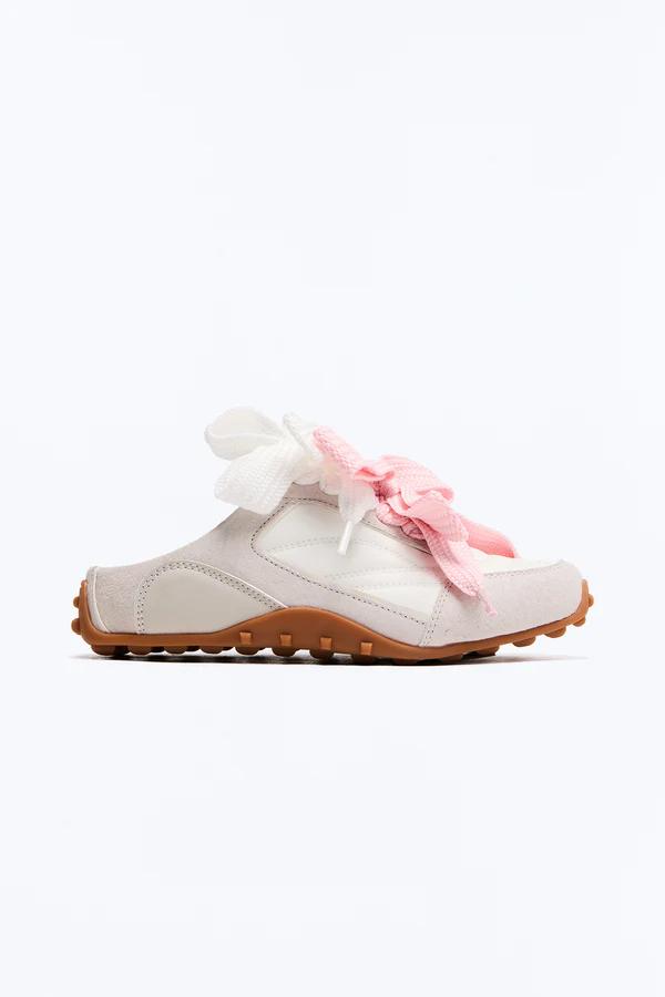 Zapatilla slip-on estilo mule, blanquecina, con detalles de gamuza beige, suela marrón texturizada y cordones rosas y blancos prominentes.