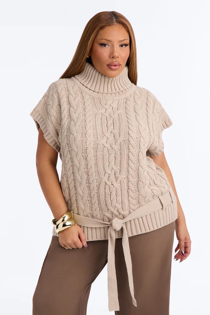 Model in beige cable-knit sleeveless turtleneck sweater vest with tie belt and brown pants.