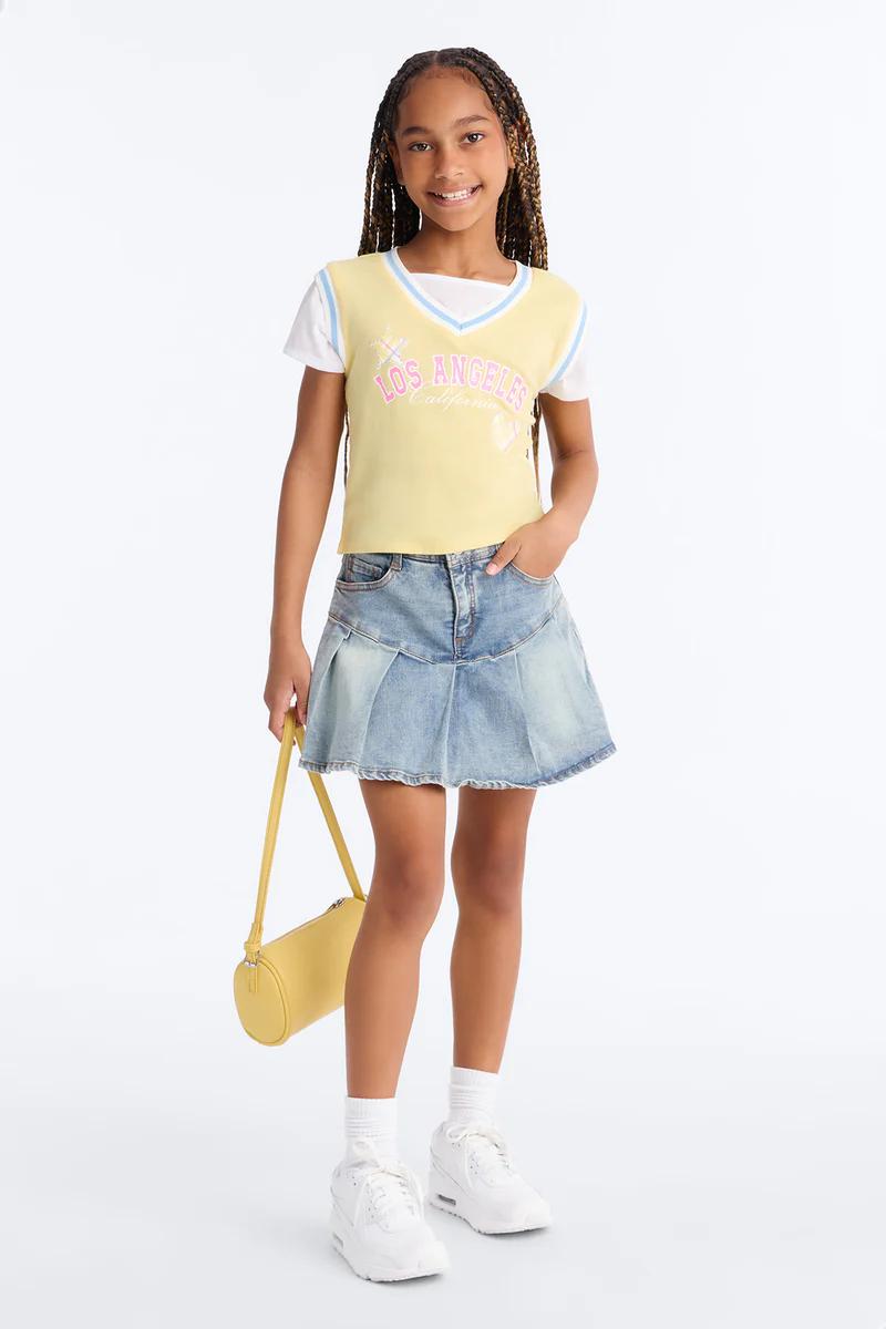 Smiling girl models yellow "Los Angeles" vest, denim pleated mini skirt, yellow bag, white sneakers. Girls' fashion outfit.