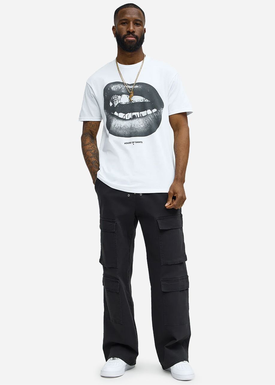 Hombre con camiseta gráfica blanca "House of Saints" de labios/grill negros, pantalones cargo negros y zapatillas blancas, mostrando moda urbana.