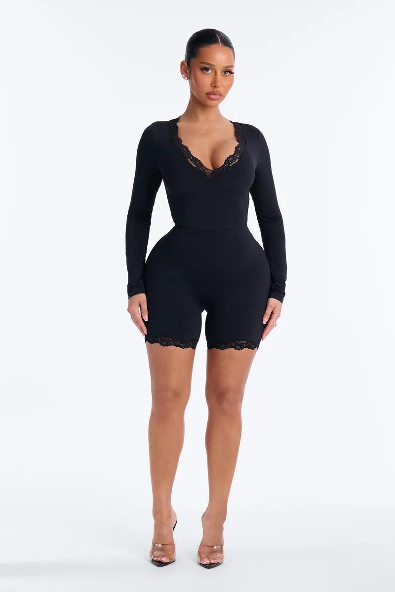 Woman wearing a black long-sleeve lace trim romper with a v-neck and fitted shorts on a plain white background.