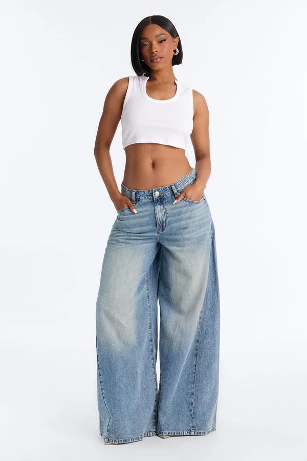 Mujer posa con top corto blanco y vaqueros de pierna ancha sobre fondo blanco, promocionando una colección de moda urbana de estilo Y2K.