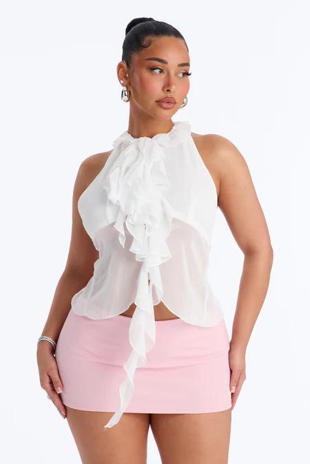 Mujer con cabello oscuro recogido, usando un top halter blanco con volantes y lazos, y una minifalda rosa. Posa sobre fondo blanco.