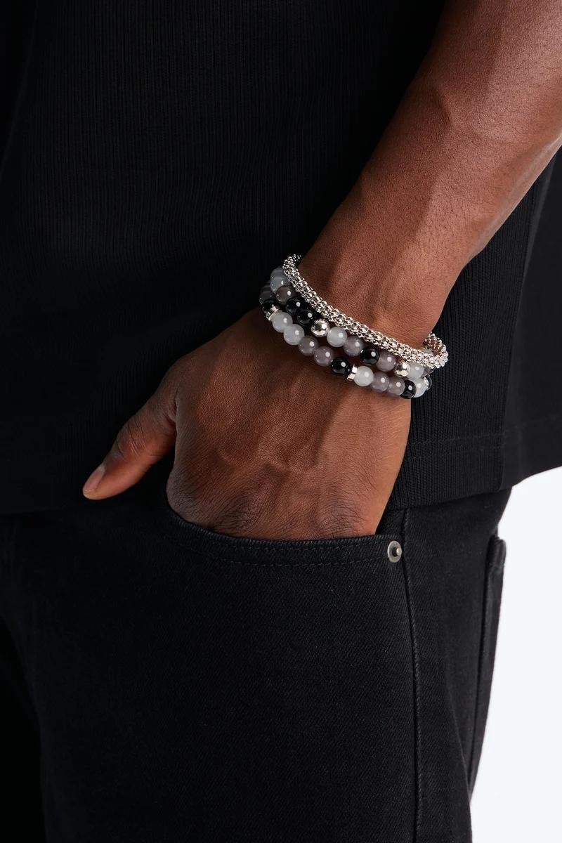 Mano en bolsillo de jeans negros mostrando pulseras apilables para hombres: cadena plateada y cuentas negras/grises. Eleva tu estilo.