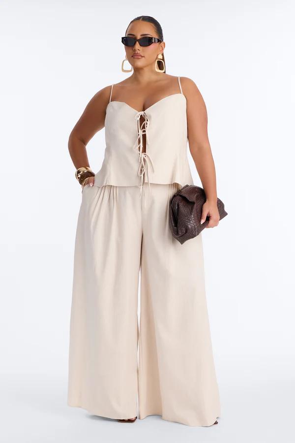 Stylish plus size woman in a beige lace-up top & wide-leg pants set, sunglasses, clutch bag. Showcasing chic summer fashion.