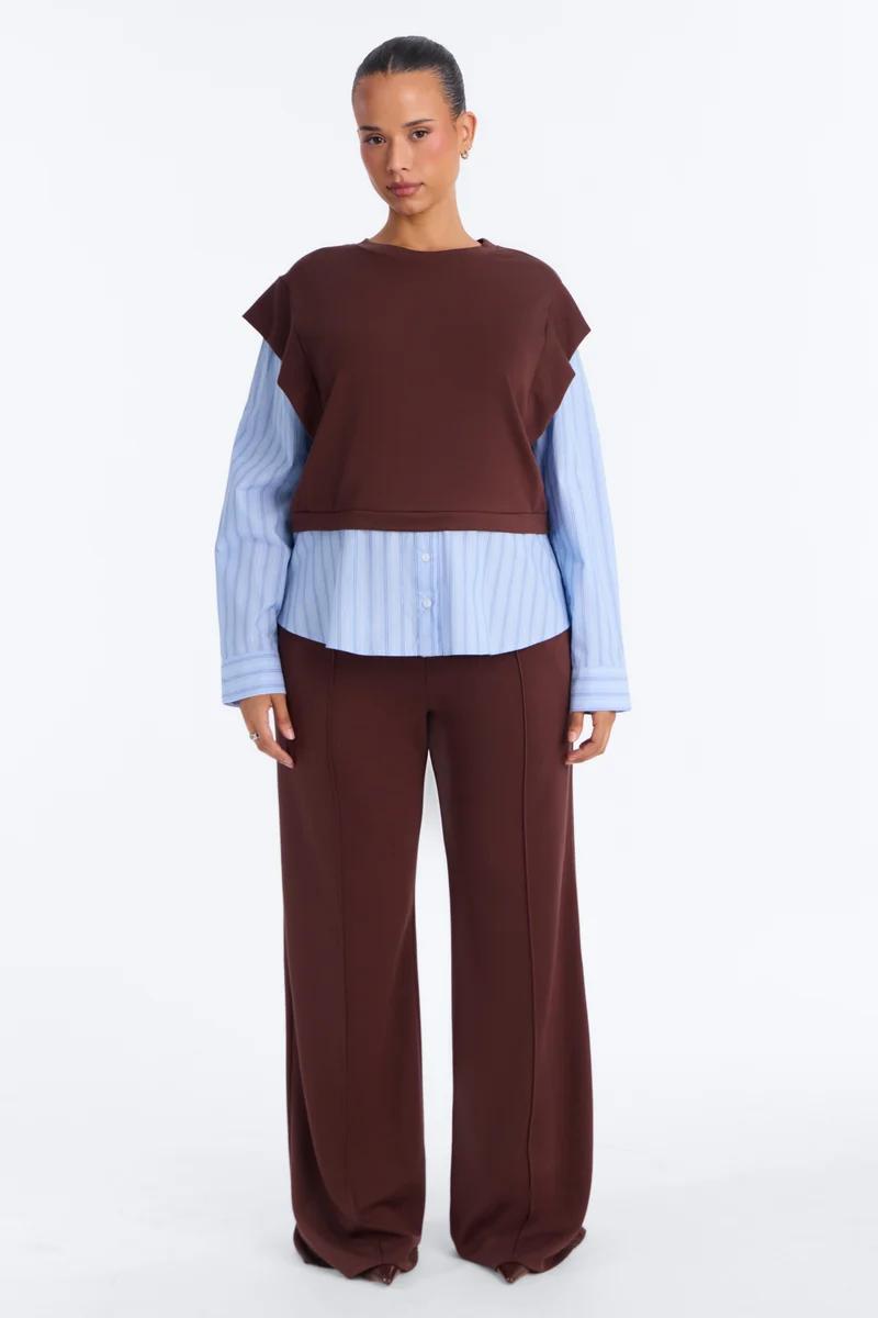 Model poses in a brown layered cropped vest over a light blue striped shirt with matching brown wide-leg pants for a modern fall collection.