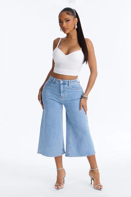 Full-body shot of a woman in a white crop top, light blue wide-leg jeans with subtle sparkles, and silver ankle-strap heels. She stands confidently.