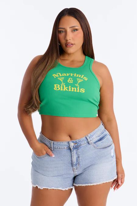 Woman in a vibrant green "Martinis & Bikinis" crop top and light wash denim shorts, posing confidently against a white background.