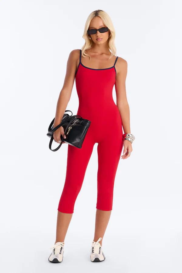 Woman in a vibrant red capri jumpsuit with black straps, stylish sunglasses, black handbag, and white sneakers, showcasing sporty fashion.