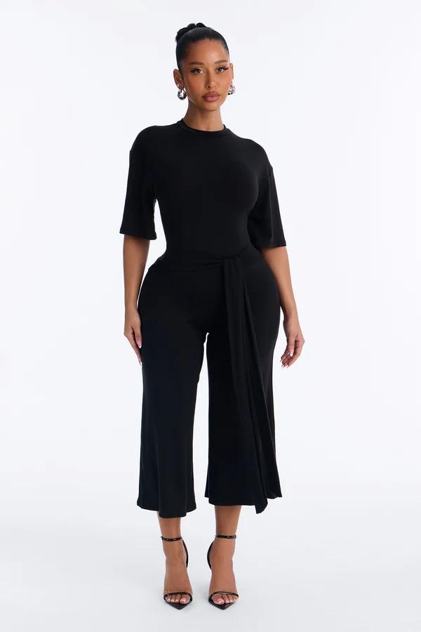 Modelo posando con un mono negro de pierna ancha, lazo en la cintura y manga corta, ideal para la nueva colección de moda casual y elegante.