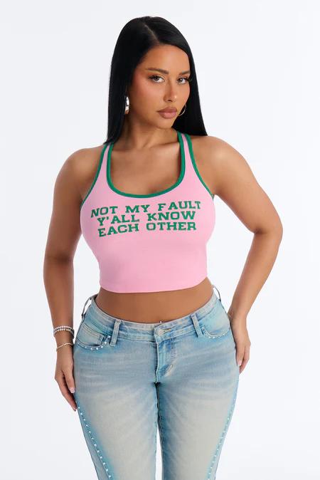 Chic woman in a pink graphic crop top with "NOT MY FAULT" text and light wash denim jeans. Promotes trendy women's apparel.