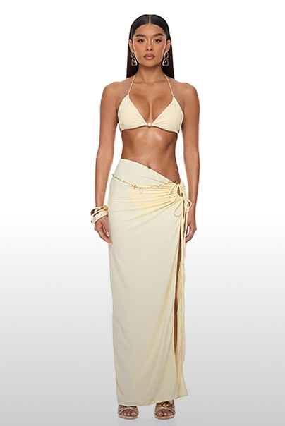 Mujer con top bikini halter crema y falda maxi fruncida con abertura, accesorios dorados. Colección ideal para resort y playa.