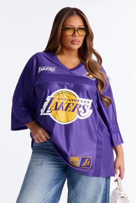 Woman wearing a purple Lakers jersey with gold logo, yellow-tinted glasses, blue jeans, and holding a white handbag.