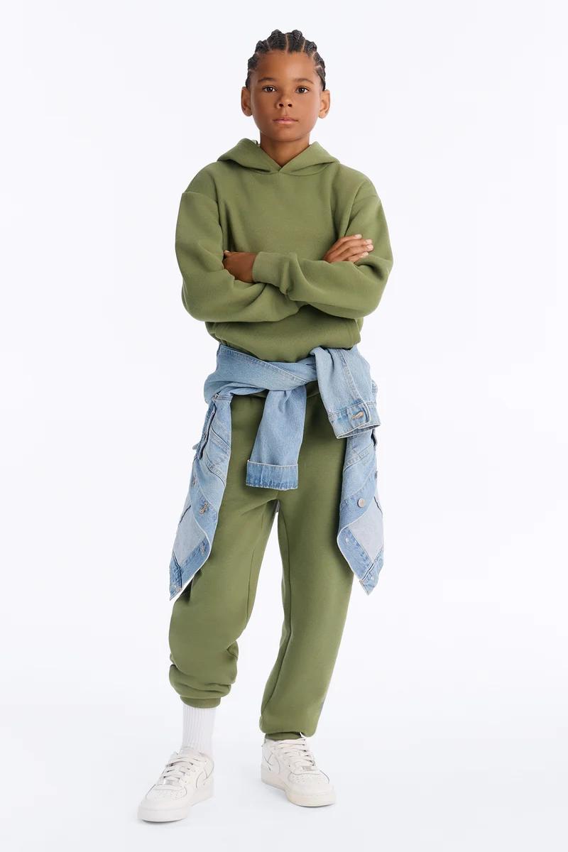 Niño con sudadera con capucha y joggers verde oliva con chaqueta de mezclilla atada a la cintura.