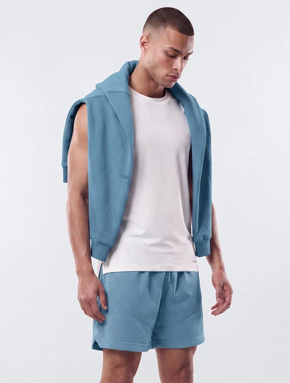 Athletic man in white tank top and blue mesh shorts with draped sweatshirt, showcasing the new men's summer activewear collection.