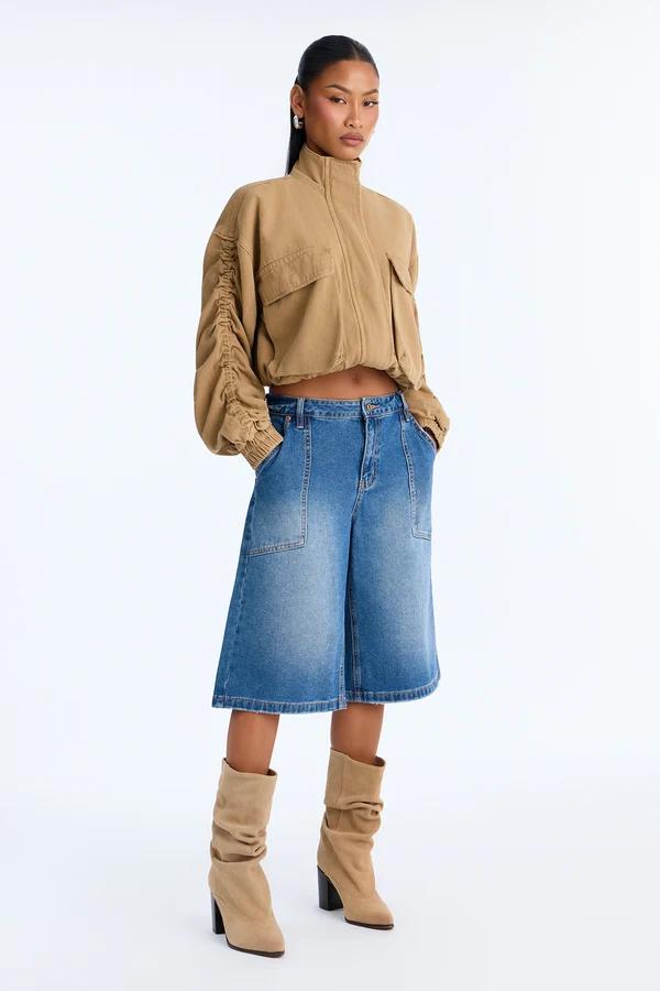 Model poses in a cropped tan utility jacket, wide-leg denim culottes, and slouchy beige boots, showcasing a trendy new streetwear collection.