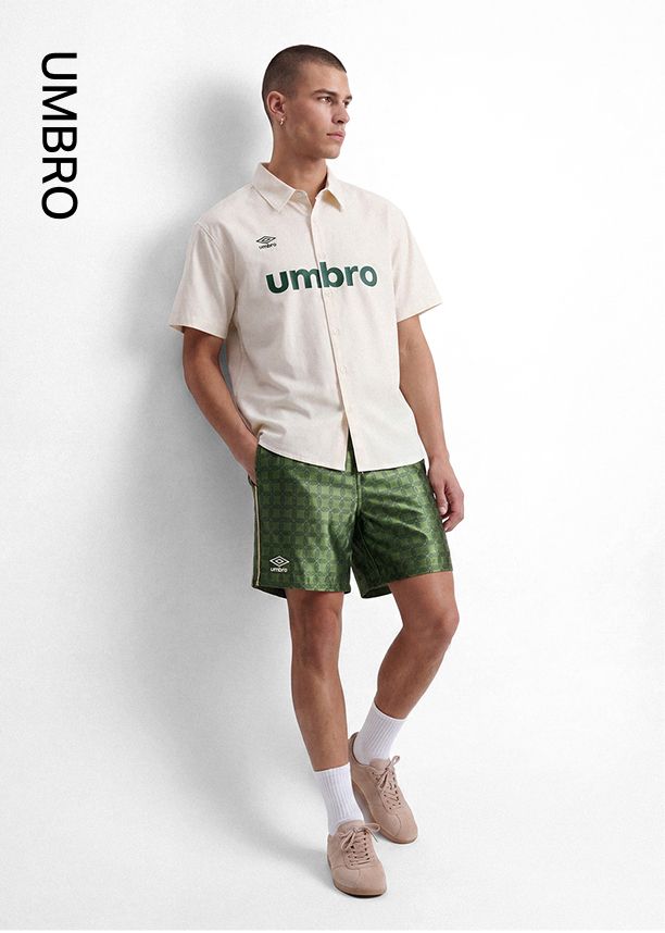 Hombre modelando camisa Umbro casual crema de manga corta con logo verde y shorts estampados verdes, promocionando moda de verano.
