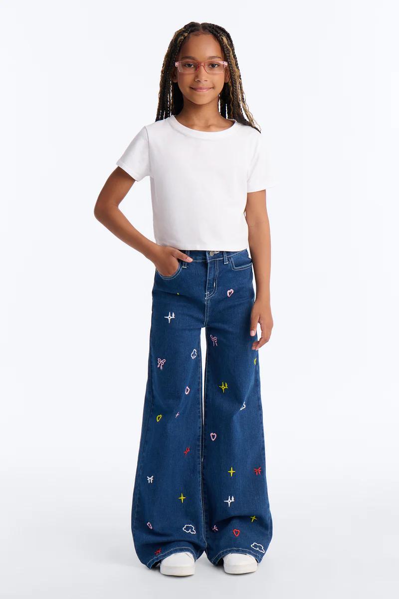 Niña con camiseta blanca corta, gafas rosas y jeans anchos azules con corazones, estrellas y lazos bordados. Moda infantil.
