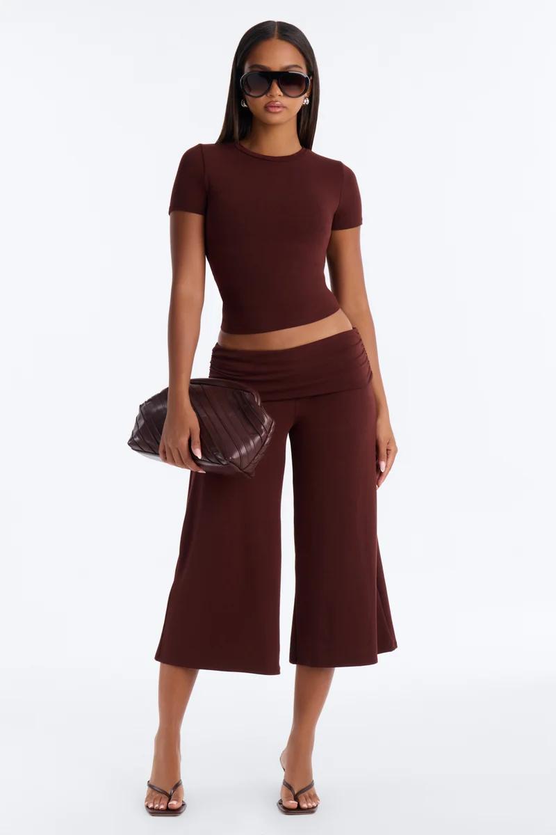 Model wears a dark brown two-piece set featuring a fitted crop top and fold-over wide-leg pants with a handbag to showcase sleek women's fashion.