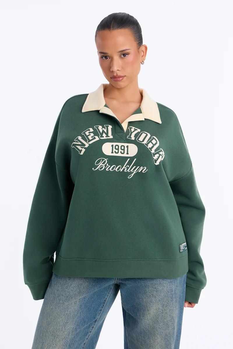 Woman wearing a dark green polo sweatshirt with New York Brooklyn graphic.